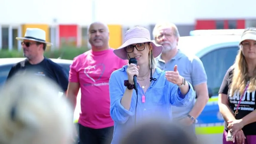 Eine Rednerin spricht bei der Kundgebung ins Mikrofon, dahinter Menschen in pinkfarbenen Team-Shirts.
